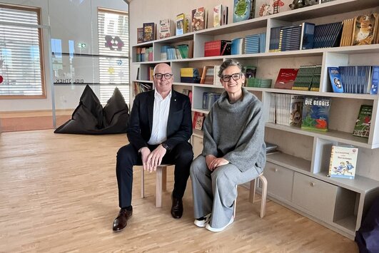 Bürgermeister Georg Rosner und Leiterin Christina Frotschnig sitzen nebeneinander auf Hockern in der Schulbibliothek.