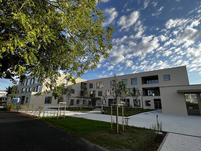 Im Vordergrund ein Ast von einem Baum mit grünen Blättern, dahinter das Gebäude der neuen Volksschule