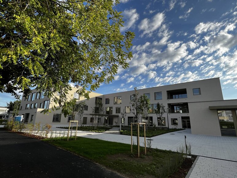 Im Vordergrund ein Ast von einem Baum mit grünen Blättern, dahinter das Gebäude der neuen Volksschule