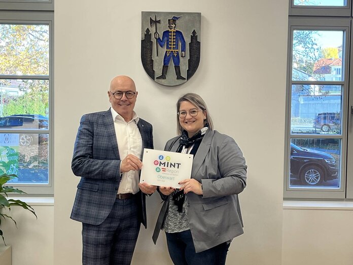 Bürgermeister und MINT-Managerin stehen im Büro des Bürgermeisters und halten die Plakette in den Händen.