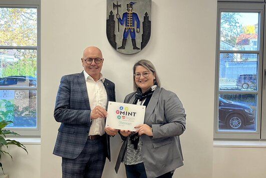 Bürgermeister und MINT-Managerin stehen im Büro des Bürgermeisters und halten die Plakette in den Händen. 