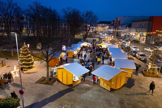 Ein Blick aus der Vogelperspektive auf den Markplatz im Stadtgarten. Es ist Abend. Die Verkaufshütten sind oval angeordnet, Menschen, die den Weihnachtsmarkt besuchen, sind am Bild zu sehen.