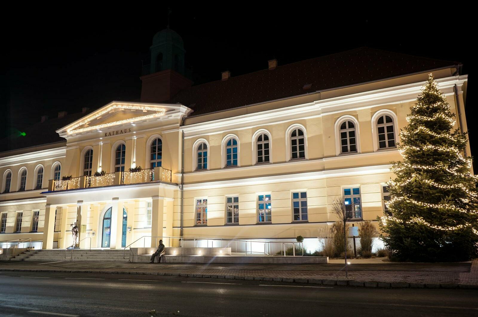 Das Rathaus in der Nacht mit weihnachtlicher Beleuchtung. Davor rechts steht ein beleuchteter Weihnachtsbaum