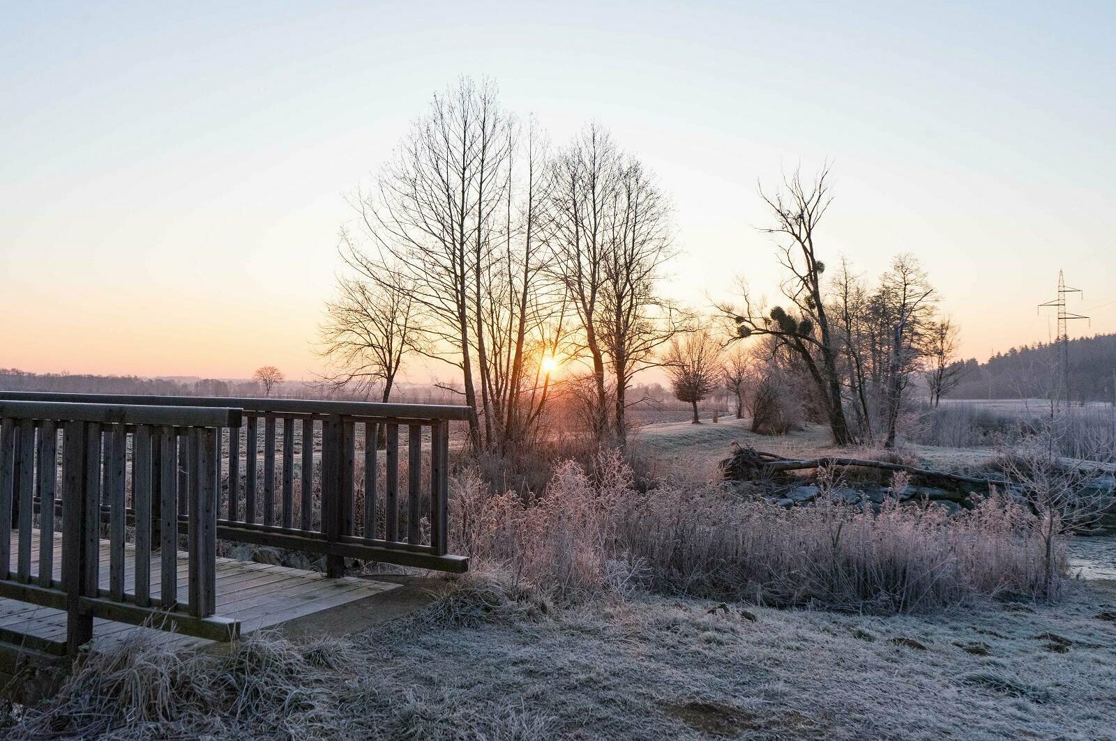 Winterliche Landschaft. Sonnenaufgang, eine leichte Schneedecke, links das Geländer einer Brücke
