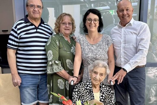 Die Jubilarin sitzt im Rollstuhl mit einem Blumenstrauß in der Hand, dahinter stehen vier Gratulanten.