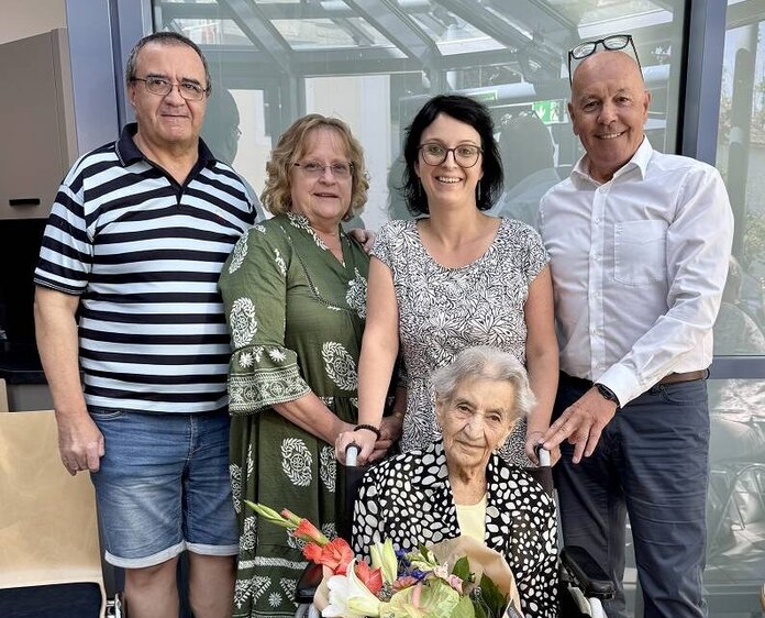 Die Jubilarin sitzt im Rollstuhl mit einem Blumenstrauß in der Hand, dahinter stehen vier Gratulanten.