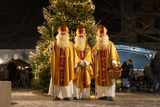 Drei Männer, die als Nikolaus verkleidet sind stehen im Stadtgarten nebeneinander, dahinter ist der beleuchtete Weihnachtsbaum zu sehen.