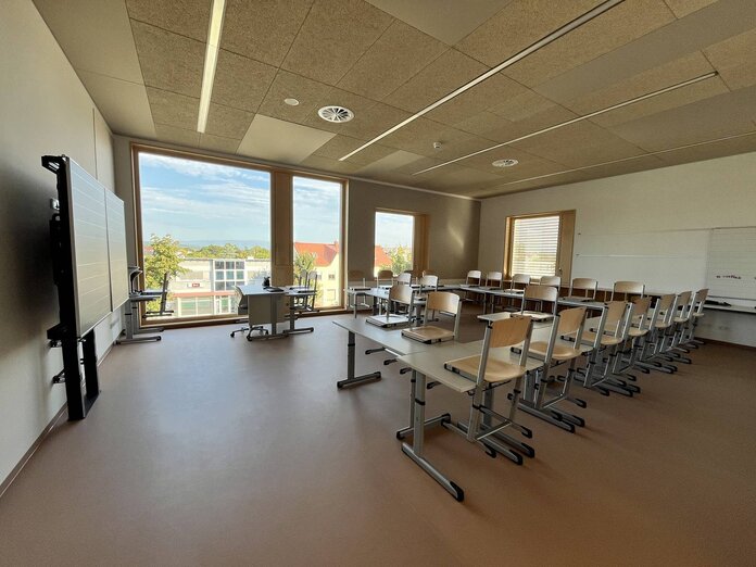 Blick in eine Klasse der neuen Volksschule, links sieht man die Tafel, rechts stehen Tische und Stühle für die Schüler. Durch eine große Fensterfront blickt man auf die Schulgasse.