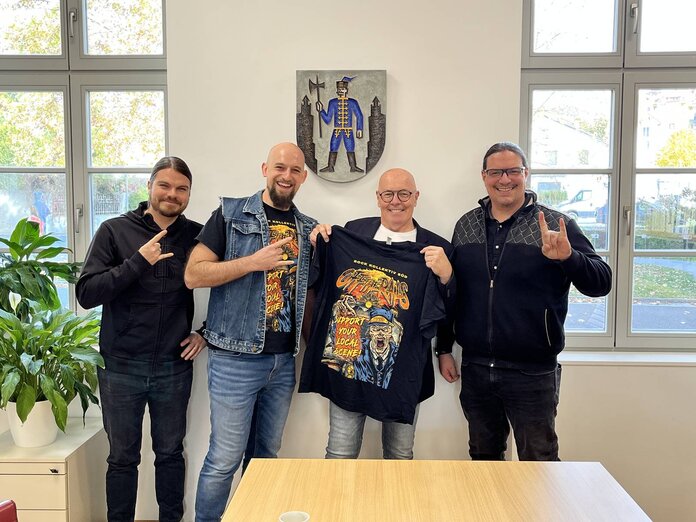 Vier Mitglieder des Rock Kollektiv Süd und der Bürgermeister stehen nebeneinander. Der Bürgermeister hält ein T-Shirt in der Hand und präsentiert es in die Kamera.