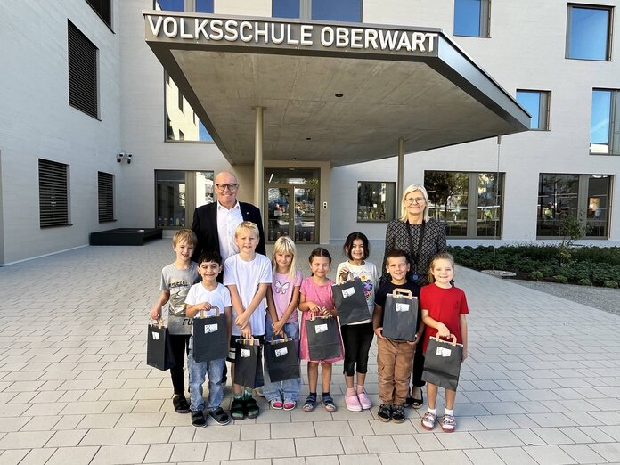 Acht Kinder, die Direktorin und der Bürgermeister stehen vor dem Eingang der neuen Volksschule. Die Kinder halten kleine Geschenkssackerl in den Händen.