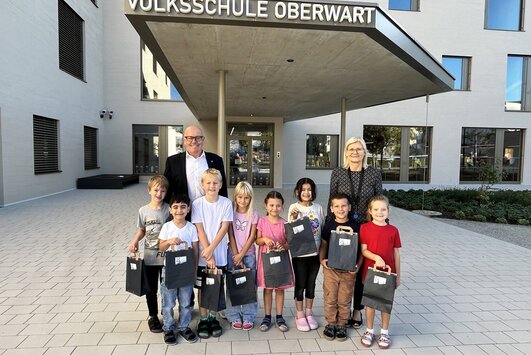 Acht Kinder, die Direktorin und der Bürgermeister stehen vor dem Eingang der neuen Volksschule. Die Kinder halten kleine Geschenkssackerl in den Händen.