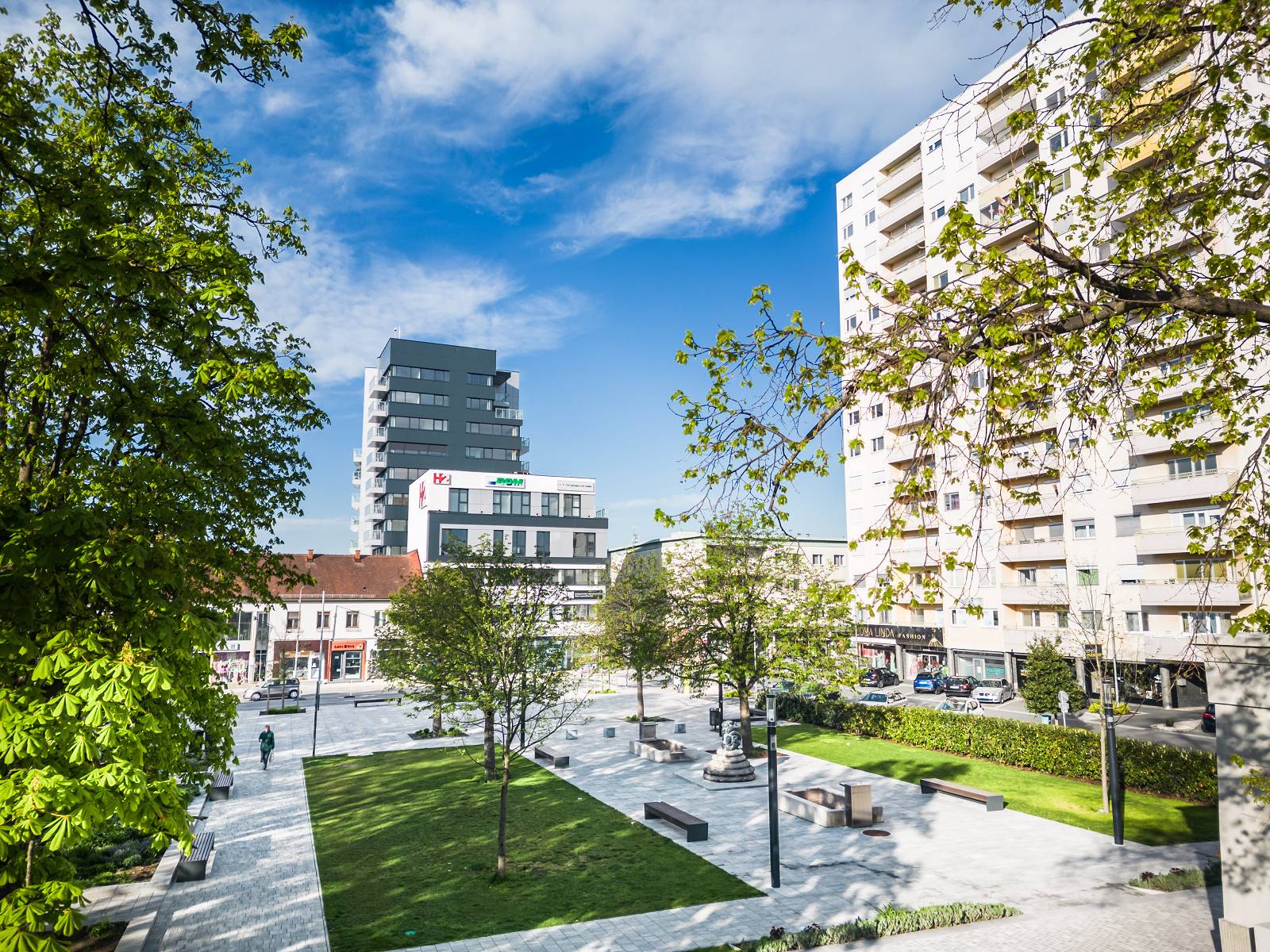 Blick in den Stadtgarten Oberwart mit Bäumen, Wegen und Sitzbänken. Im Hintergrund das Hochhaus und der H2-Tower.