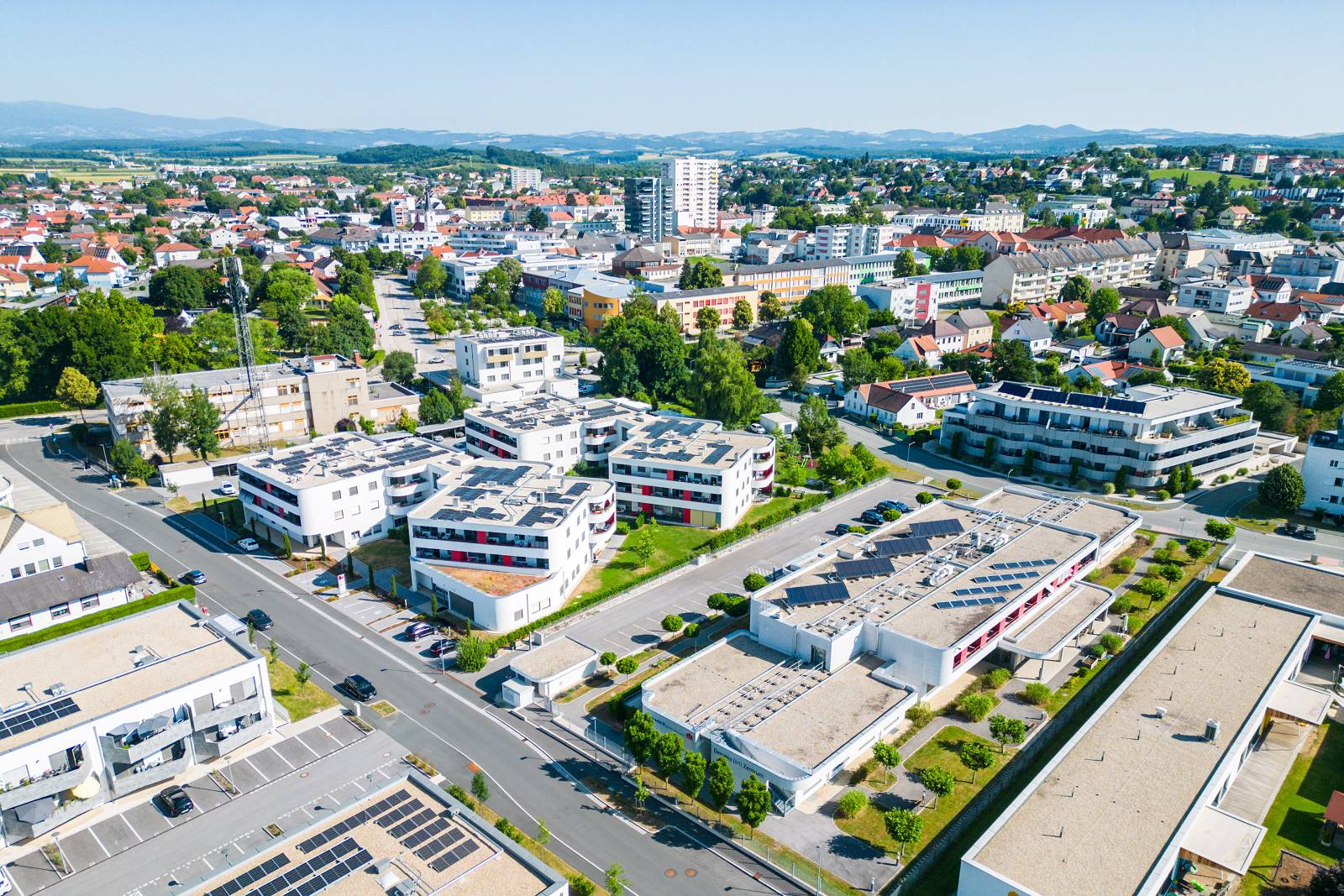 Luftbild der Stadtgemeinde Oberwart an einem sonnigen Tag. Im Vordergrund moderne Wohnbauten mit Flachdächern und Solaranlagen, daneben ein Kindergartengebäude mit hellen Fassaden und begrüntem Umfeld. Dahinter dicht bebaute Innenstadt mit einer Mischung aus Einfamilienhäusern, mehrstöckigen Wohngebäuden und dem markanten Hochhaus. Im Hintergrund hügelige Landschaft mit Wäldern und Feldern. Die Straßen sind ruhig, einige Autos sind geparkt.